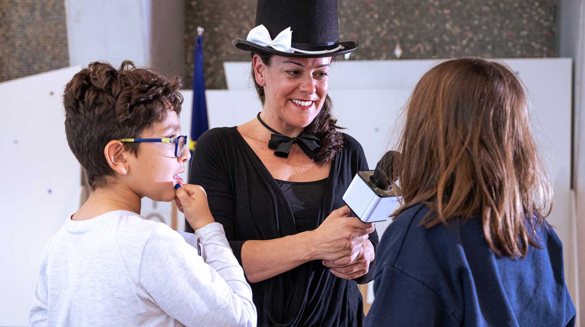 Nazareth wearing a toll hat with a white ribbon is holding a mic and interviewing two young people.